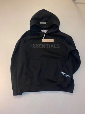 Svart Essentials hoodie Fear of God - Snygg svart hoodie från Fear of God Essentials med stor ESSENTIALS-text på bröstet och mindre logga på ärmen. Klassisk känguruficka framtill och huva. Tillverkad i mjukt bomullsmaterial som känns riktigt skönt mot huden. Perfekt för en clean och avslappnad streetwear-look.