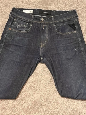 Mörkblå jeans från Replay - Snygga mörkblå jeans från Replay med klassisk femficksmodell och kontrastsömmar. Jeansen har diskreta slitningar vid fickorna och en rak passform. Tillverkade i denim med detaljer i silverfärgad metall. Perfekta för en trendig och avslappnad stil. Waist 29 Lengt 32