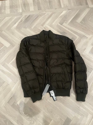 Mörkgrön pufferjacka från J Lindeberg  - Snygg mörkgrön pufferjacka från J Lindeberg. Den är omvändbar så ena sidan funkar som puffer och andra mer som bomberjacka. Den är alldrig andvänd. Skriv om du har några frågor. 