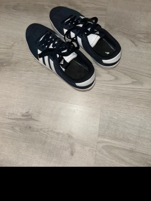 Adidas Originals mörkblå/vita sneakers - Snygga Adidas Originals sneakers i mörkblå med klassiska vita ränder på sidorna och vit sula. Skorna har lågt skaft och är tillverkade i mockaliknande material med textildetaljer. Perfekta för en sportig och stilren look.