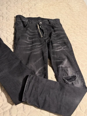 Svarta dsquared2 jeans - Säljer ett par svarta dsquared2 jeans med slitningar  Jeansen har en cool dragkedjedetalj vid gylfen och klassisk femficksmodell. Materialet är denim med stretch för skön passform. Perfekta för dig som gillar en edgy och avslappnad stil.