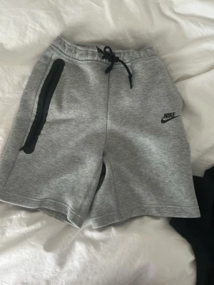Grå Nike tech shorts - Snygga gråa sweatshorts från Nike med svart logga på vänster ben och svarta detaljer vid fickorna. Shortsen har snörning i midjan och är tillverkade i mjuk bomullsblandning, perfekta för chill dagar eller träning.