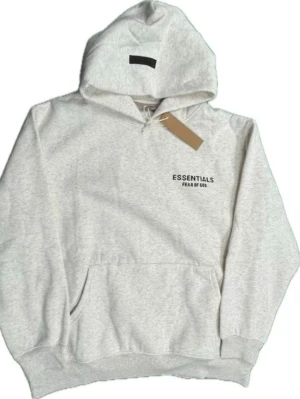 Essentials Fear of God hoodie - Snygg ljusgrå hoodie från Essentials Fear of God med svart logga tryckt på bröstet och ryggen. Klassisk känguruficka framtill, ribbade muddar och huva med dragsko. Tillverkad i mjukt bomullsmaterial som är perfekt för en avslappnad streetwear-look.