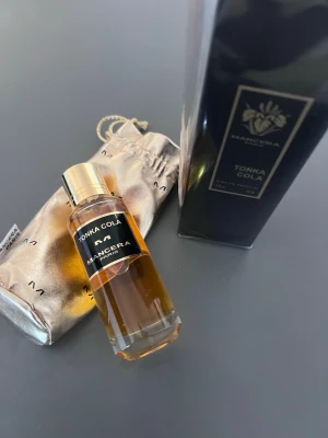 Mancera Tonka Cola Eau de Parfum - Mancera Tonka Cola Eau de Parfum med elegant glasflaska, guldfärgat lock och svart etikett. Doften kommer i en lyxig svart förpackning och med en skimrande guldpåse. Perfekt för dig som gillar unika och djupa doftnoter med tonkaböna och cola.