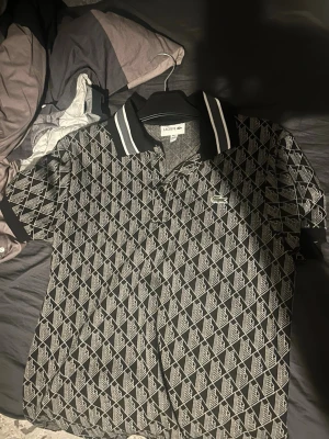 Svart mönstrad skjorta från Lacoste - Säljer en svart skjorta från Lacoste med vitt geometriskt mönster och klassisk krage med vita ränder. Skjortan har korta ärmar, knappar framtill och den ikoniska gröna krokodilloggan på bröstet. Perfekt för dig som vill sticka ut med en unik stil.