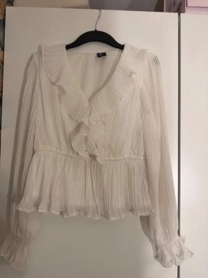 Vit omlottklänning med volang - Säljer en vit kort omlottklänning med volangdetaljer och knyt i midjan. Klänningen har långa ärmar med resår vid ärmslut och en v-ringad halsringning. Perfekt för dig som gillar en feminin och trendig stil. Har däremot sminfläckar på in sidan men syns inte! 