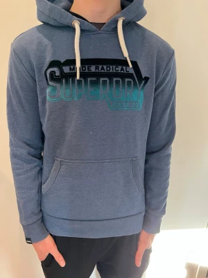 Blå Superdry hoodie med tryck - Snygg blå hoodie från Superdry i storlek S modellen är 181cm och väger 66kg 