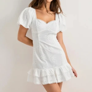 Nelly studentklänning - Söt vit kortklänning med puffärmar, volangkant nedtill och broderade blommor. Klänningen har fyrkantig ringning och dekorativa spetsdetaljer längs kanterna. Perfekt för sommarens alla tillfällen. Helt ny med lapp kvar