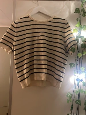 Randig kortärmad topp från H&M - Säljer en krämvit kortärmad topp från H&M med svarta horisontella ränder. Toppen har rund halsringning och är stickad i mjukt material. Perfekt för en avslappnad och trendig look.