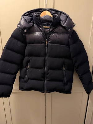 Pufferjacka från Polo Ralph Lauren - Snygg mörkblå pufferjacka från Polo Ralph Lauren med glansiga partier upptill och på huvan. Jackan har dragkedja framtill, två fickor med dragkedja och broderad logga på bröstet. Perfekt för kalla vinterdagar och riktigt stilren look. Finns ett minimalt hål i jackan (bild 2) inget som syns. Den är även lagad i luvan men heller inget man tänker på (bild 4). Hör av er vid frågor.