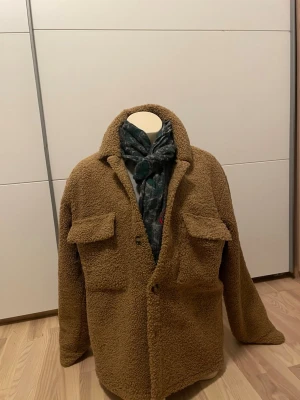 Brun teddyjacka med fickor - Mysig brun teddyjacka i oversized modell med två stora bröstfickor och knäppning framtill. Jackan har klassisk krage och är gjord i fluffigt teddy-material som ger en trendig look. Perfekt för dig som vill ha en varm och stilren jacka till hösten eller vintern.