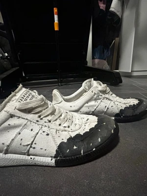 Maison Margiela vita sneakers med svart tå - Maison Margiela sneakers i vitt tyg med distinkt svart tåparti och färgstänk. Klassisk låg modell med vita snören och märkets ikoniska etikett på plösen. Snyggt slitna detaljer och unik design som sticker ut. Perfekta för dig som gillar statement-plagg. Använda ett par fåtal gånger så priset är inte helt fast. Hör gärna av er vid intresse!