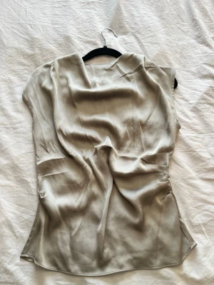 Beige ärmlös sidenblus - Stilren beige blus i siden med ärmlös design Toppen har en figurnära passform och dragkedja i ryggen. 