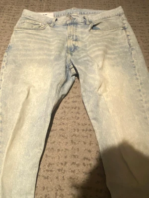 Ljusblå jeans - Säljer ett par ljusblå slim fit jeans med klassisk femficksmodell och dragkedjegylf. Jeansen har en snygg tvättad look med slitningar framtill och smal passform som sitter tajt längs benen. Perfekta för en avslappnad och trendig stil.