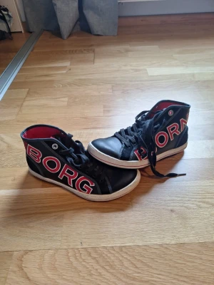 Svarta Björn Borg sneakers med rosa inslag - Snygga svarta sneakers från Björn Borg med stora rosa bokstäver på sidan. Har en klassisk high-top design som ger en cool streetstil.