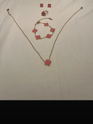 Smyckesset med rosa fyrklöver - Säljer ett smyckesset med halsband, armband, ring och örhängen i guldton med rosa fyrklöverhängen. Setet har en elegant och trendig design med detaljerade kanter runt varje hänge. Perfekt för dig som gillar färgglada accessoarer och vill sticka ut.