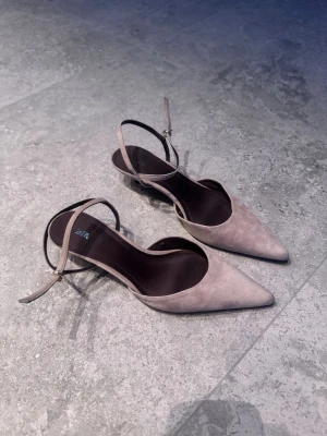 Beige spetsiga pumps från Zara - Säljer ett par snygga pumps från Zara i beige mocka med spetsig tå och smal ankelrem med guldfärgat spänne. Skorna har en elegant och modern design med öppen sida och mörk innersula. Perfekta för dig som vill ha en stilren look.