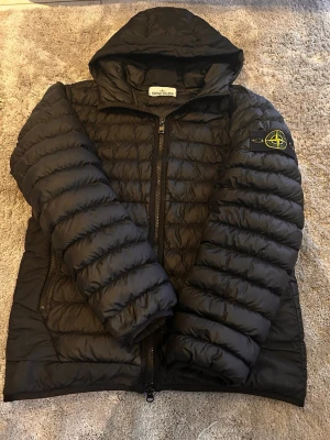 Svart pufferjacka från Stone Island - Snygg svart pufferjacka från Stone Island med huva och dragkedja framtill. Jackan har det klassiska Stone Island-märket på vänster ärm och två sidofickor med dragkedja. Tillverkad i lätt, quiltat material som håller värmen under kalla dagar.