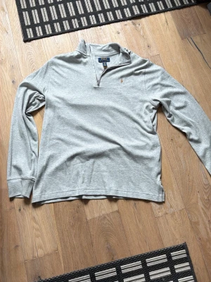 Grå långärmad tröja från Polo Ralph Lauren - Säljer en stilren grå långärmad tröja från Polo Ralph Lauren med klassisk halvzip och broderad logga på bröstet. Tröjan har en mjuk bomullskänsla och passar perfekt till både jeans och chinos. Enkel och tidlös design med skön look. Aldrig använd!