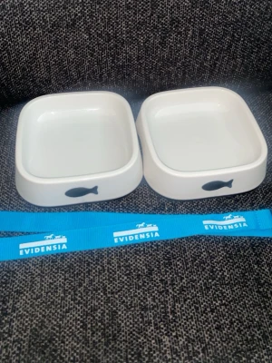 Två vita matskålar för husdjur - Två vita fyrkantiga matskålar i keramik med svart fiskmotiv på framsidan. Perfekta för katt eller liten hund. Medföljer gör ett blått band med texten 'Evidensia'.