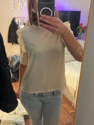 Beige blus med volangärmar - Säljer en beige blus med korta ärmar och volangdetaljer på axlarna. Blusen är lätt transparent och har en rak passform som ger en avslappnad look. Perfekt att styla med jeans eller kjol för en trendig outfit.