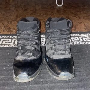 Svarta höga sneakers med glansig finish - Säljer ett par svarta höga sneakers från Jordan med glansig finish på tå och sidor, samt matta partier i textil. Skorna har kraftiga svarta snören och en robust sula som ger en sportig vibe. Perfekta för dig som gillar klassisk streetwear-stil.