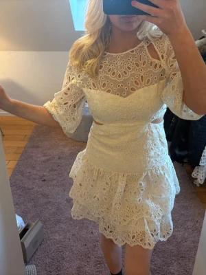 Vit spetsklänning med volanger - Säljer en superfin vit kortklänning med broderad spets, volangdetaljer och trekvartsärm. Klänningen har en omlottkänsla upptill, markerad midja och vackra hålbroderier som ger en romantisk look. Perfekt för sommarens alla tillfällen! Benvit färg