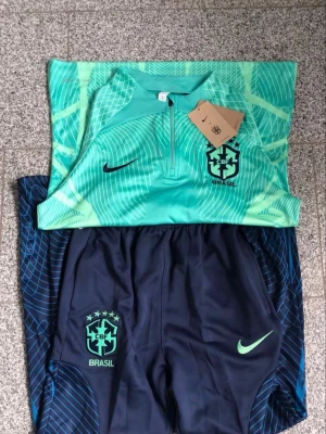 Nike Brasil träningsset långärmad - Snyggt träningsset från Nike med Brasil-logga. Överdelen är långärmad i turkos med grafiskt mönster och broderad CBF-logga. Byxorna är mörkblå med turkosa detaljer och Nike-logga. Tillverkat i lätt, snabbtorkande Dri-FIT-material som andas.