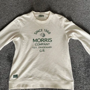 Vit Morris tröja  - Vit tröja från Morris med grönt tryck framtill där det står 'Since 1968 Morris Company Est. Trademark G.B.' och ett lejonhuvud. Klassisk rund halsringning och ribbade muddar vid ärmslut och nederkant. Perfekt för en clean och stilren look. Passar perfekt inför vår och sommar.
