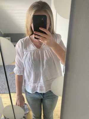 Vit boho-blus med spetsdetaljer - Luftig vit blus med v-ringning och fina spetsdetaljer längs bröstet och ärmsluten. Blusen har trekvartsärmar med lätt volang. Storlek 164