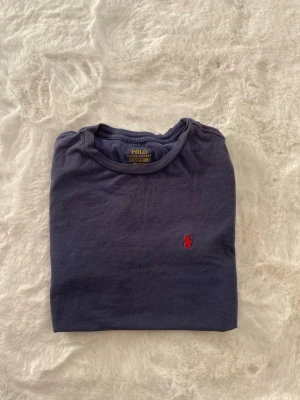 Blå t-shirt från Polo Ralph Lauren - Säljer en klassisk blå t-shirt från Polo Ralph Lauren med det ikoniska röda broderade logotypen på bröstet. Perfekt för en stilren och avslappnad look. Tillverkad i mjukt material för extra komfort.