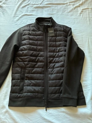 Svart dunväst från Dressmann - Helt oanvänd. Pristagen är kvar. Snygg svart dunväst från Dressmann med quiltad framsida och släta ärmar. 
