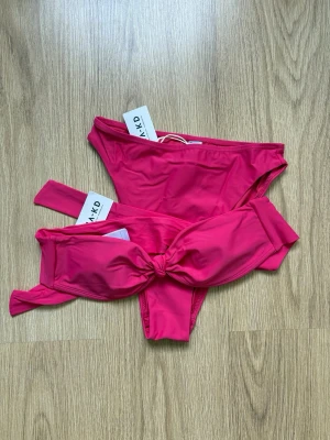 Rosa bikini med knut från NA-KD - Snygg rosa bikini från NA-KD i storlek XS. Överdelen har en trendig knut framtill, medan underdelen har en hög skärning för en modern look. Perfekt för strandhäng eller poolparty. Materialet är mjukt och stretchigt