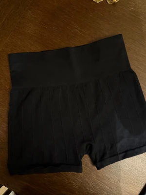 Svarta högmidjade shorts från H&M Sport - Säljer ett par svarta högmidjade shorts från H&M Sport. De är ribbade och har en tight passform med bred linning för extra stöd. Perfekta för träning eller chill dagar. Materialet är mjukt och stretchigt syntetmaterial.