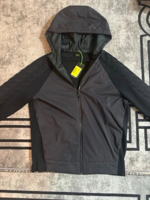 Marinblå cardigan med huva från BOSS - Stilren svart vindjacka från BOSS med hel dragkedja och huva. Jackan har diskret BOSS-logga på bröstet, mönstrade svarta ärmar och sportig passform. Perfekt för blåsiga dagar och enkel att matcha med olika outfits. Ordinarie priset var 2200kr 