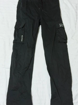 Svarta cargobyxor med fickor - Säljer ett par svarta cargobyxor med stora sidofickor och dragsko i midjan. Byxorna har tryckdetaljer på fickorna och är tillverkade i ett mjukt, slitstarkt tyg. Perfekta för en avslappnad streetwear-look.