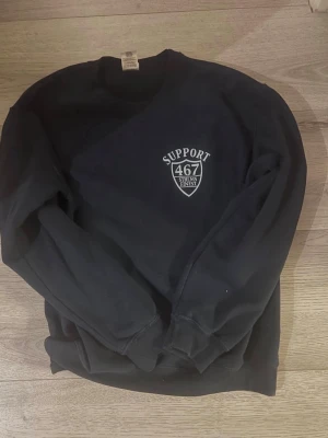 Svart sweatshirt SUPPORT 467 - Cool svart sweatshirt med vitt SUPPORT 467 STHLMS FINEST-tryck både fram och stort på ryggen. Klassisk rund hals och ribbade muddar vid ärmar och nederkant. Perfekt för dig som gillar streetwear och vill sticka ut.