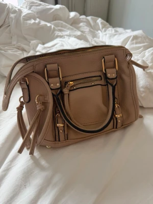 Beige handväska med gulddetaljer - Snygg beige handväska i mjukt skinnliknande material med guldfärgade metalldetaljer och dragkedjor. Köpt i Italien för 30 euro, aldrig använd!