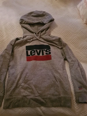 Grå Levi's hoodie med logga - Snygg grå hoodie från Levi's med stor logga i mörkblå, vitt och rött framtill. Klassisk modell med huva och dragsko samt magficka. Tillverkad i mjukt bomullsmaterial, perfekt för en avslappnad streetstyle-look.