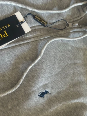Grå mjukisbyxor från Polo Ralph Lauren - Snygga grå mjukisbyxor från Polo Ralph Lauren med klassisk broderad logga i mörkblått på benet. Byxorna har dragsko i midjan och ribbade muddar vid bensluten. Perfekta för en avslappnad och stilren look.