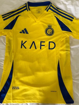Gul och blå Al Nassr matchtröja Adidas - Snygg gul matchtröja från Al Nassr FC med blå detaljer och klubbmärke på bröstet. Tillverkad i lätt och ventilerande polyester med Aeroready-teknologi. Klassisk Adidas-design med tre ränder på axlarna och tryckta loggor framtill.