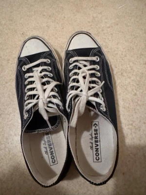 Svarta Converse Chuck Taylor sneakers - Klassiska svarta Converse Chuck Taylor sneakers med vit tå och vita snören. Lågt skaft, vit sula med svarta ränder och tydliga kontrastsömmar. Ovandel i canvas och innerfoder i textil. Perfekta för en avslappnad streetstil.