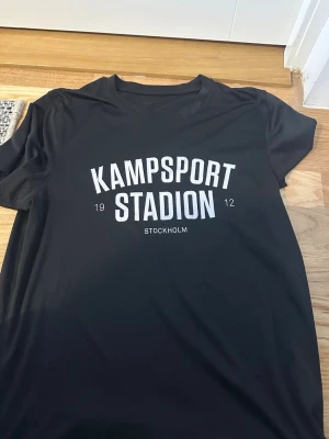 Tränings  t-shirt Kampsport Stadion - Svart t-shirt med vit text 'Kampsport Stadion 1912 Stockholm' tryckt på bröstet. Klassisk rund hals och korta ärmar, perfekt för en sportig och avslappnad stil. Material: polyester 