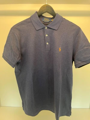 Blå pikétröja från Polo Ralph Lauren - Klassisk blå pikétröja från Polo Ralph Lauren med slim fit-passform. Tröjan har korta ärmar, krage och tre knappar framtill samt den ikoniska broderade loggan på bröstet. Perfekt för en stilren och avslappnad look.