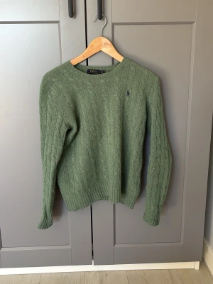Grön kabelstickad tröja Polo Ralph Lauren - Kabelstickad grön tröja från Polo Ralph Lauren med rund halsringning och broderad logga på bröstet. Tröjan har långa ärmar och ribbade muddar vid ärmslut och nederkant. Perfekt för lager-på-lager och snygg till jeans.