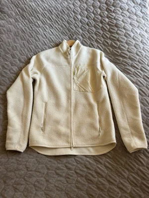 Peak Performance fleecejacka - Mysig beige fleecejacka med hel dragkedja framtill, hög krage och tre fickor med dragkedja. Jackan har en bröstficka i nylonmaterial och tumhål vid ärmsluten. Köptes sommaren 2025 och har i princip bara hängt i garderoben. Använd enstaka gånger. 