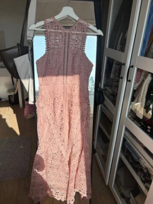 Rosa spetsklänning med hög hals - Superfin rosa långklänning i spets med hög hals och ärmlös design. Klänningen har vackra broderade detaljer och en feminin siluett. Perfekt för dig som vill sticka ut med en romantisk look. Materialet känns luftigt och elegant. Har en väldigt fin och figurnära passform. 