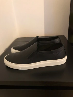 Svarta slip-on sneakers från Kavat - Stilrena svarta slip-on sneakers från Kavat med ovandel i slätt läder och vit sula. Skorna har elastiska paneler på sidorna för enkel påtagning och en klassisk låg profil. Perfekta för en clean och modern look.