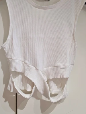 Vit ribbad linnetopp från Pull&Bear - Snygg vit ribbad linnetopp från Pull&Bear med coola utskurna detaljer framtill. Toppen är ärmlös och har en croppad passform som ger en trendig look. Perfekt att matcha med jeans eller kjol för en stilren outfit.