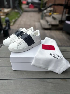 Valentino Garavani vita sneakers - Snygga vita sneakers från Valentino Garavani med en bred svart rem över mitten. Klassisk låg modell med vita snören och gummisula. Tillverkade i läder för en lyxig känsla och stilren look. Perfekta för dig som vill sticka ut med exklusiva detaljer.